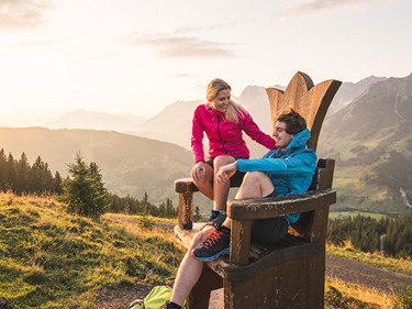 Ein Pärchen sitz auf einem Holzstuhl mit Kronendetail und blicken hinab auf das Tal bei Sonnenaufgang.